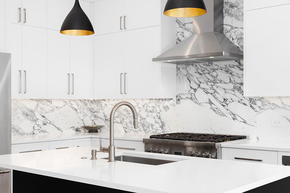 A kitchen detail with white cabinets, modern light fixtures hanging over a black island, and a marble countertop and backsplash.