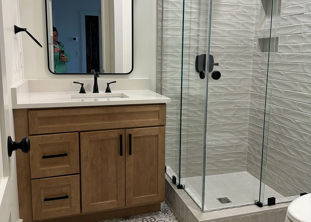 Bathroom remodel with a natural wood vanity, black fixtures, and a textured tile walk-in shower.