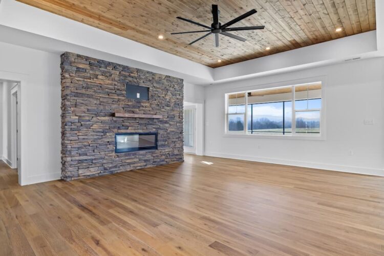 Living room with stacked stone fireplace, wood mantel, wood plank ceiling, black ceiling fan, hardwood floors, and large window with mountain views