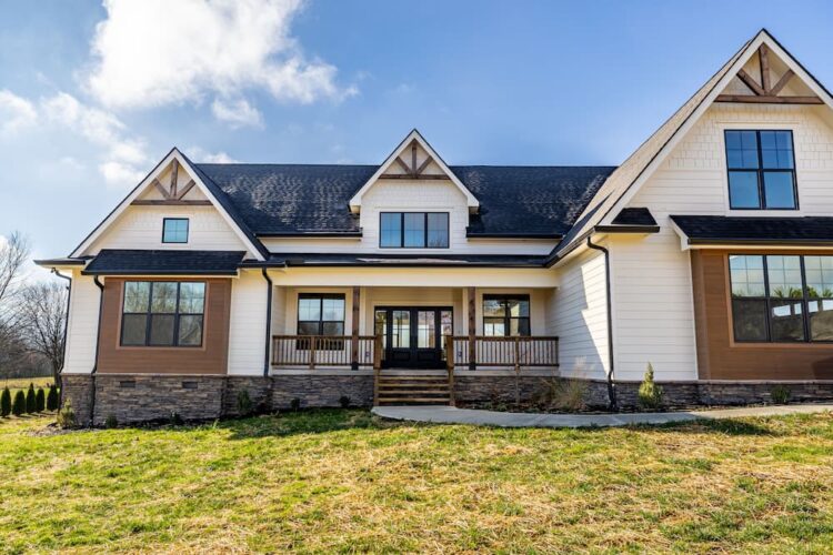 Front exterior of modern farmhouse with white siding, brown accent panels, timber gable brackets, covered porch with black French doors, and stacked stone foundation